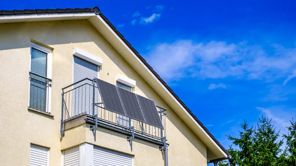 Drei schwarze Solarmodule eines Balkonkraftwerks, die mit einer stabilen Halterung an einem Metall-Gitterbalkon eines gelben Einfamilienhauses montiert sind.